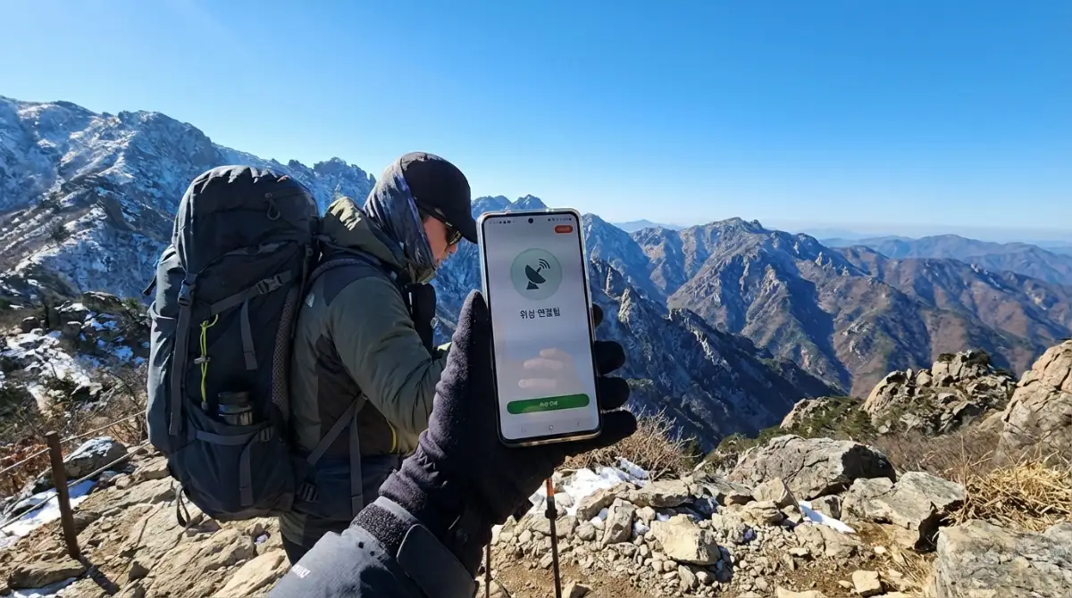 맑은 파란 하늘 아래 깊은 산맥에서 갤럭시 S26 울트라의 위성 연결 아이콘을 확인하며 등산하는 고화질 이미지
